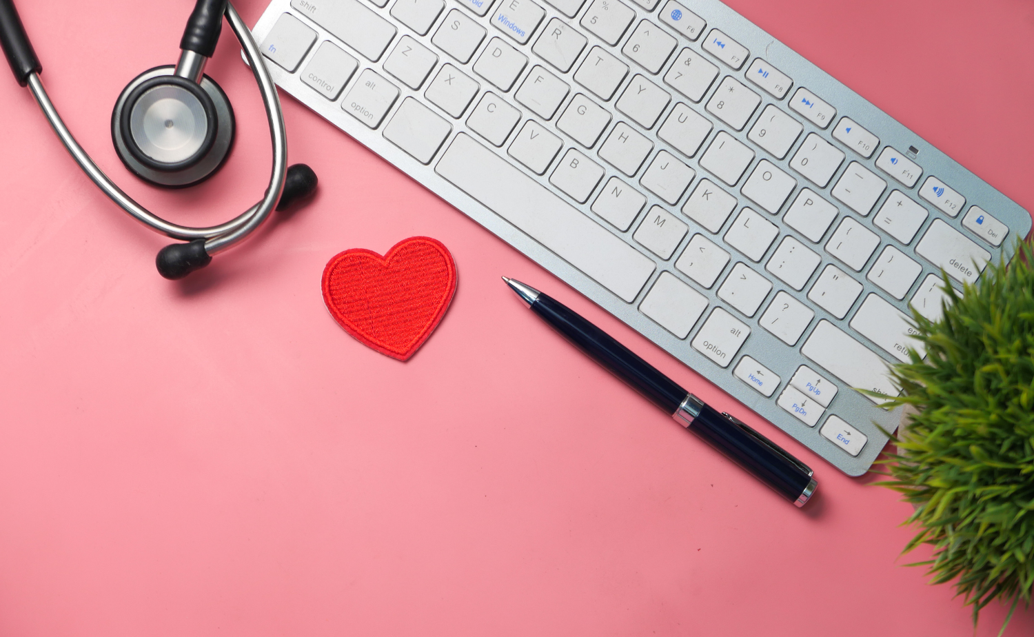 A stethoscope, a red heart, a pen, and a keyboard are arranged on a pink background, symbolizing the intersection of healthcare, technology, and compassion.