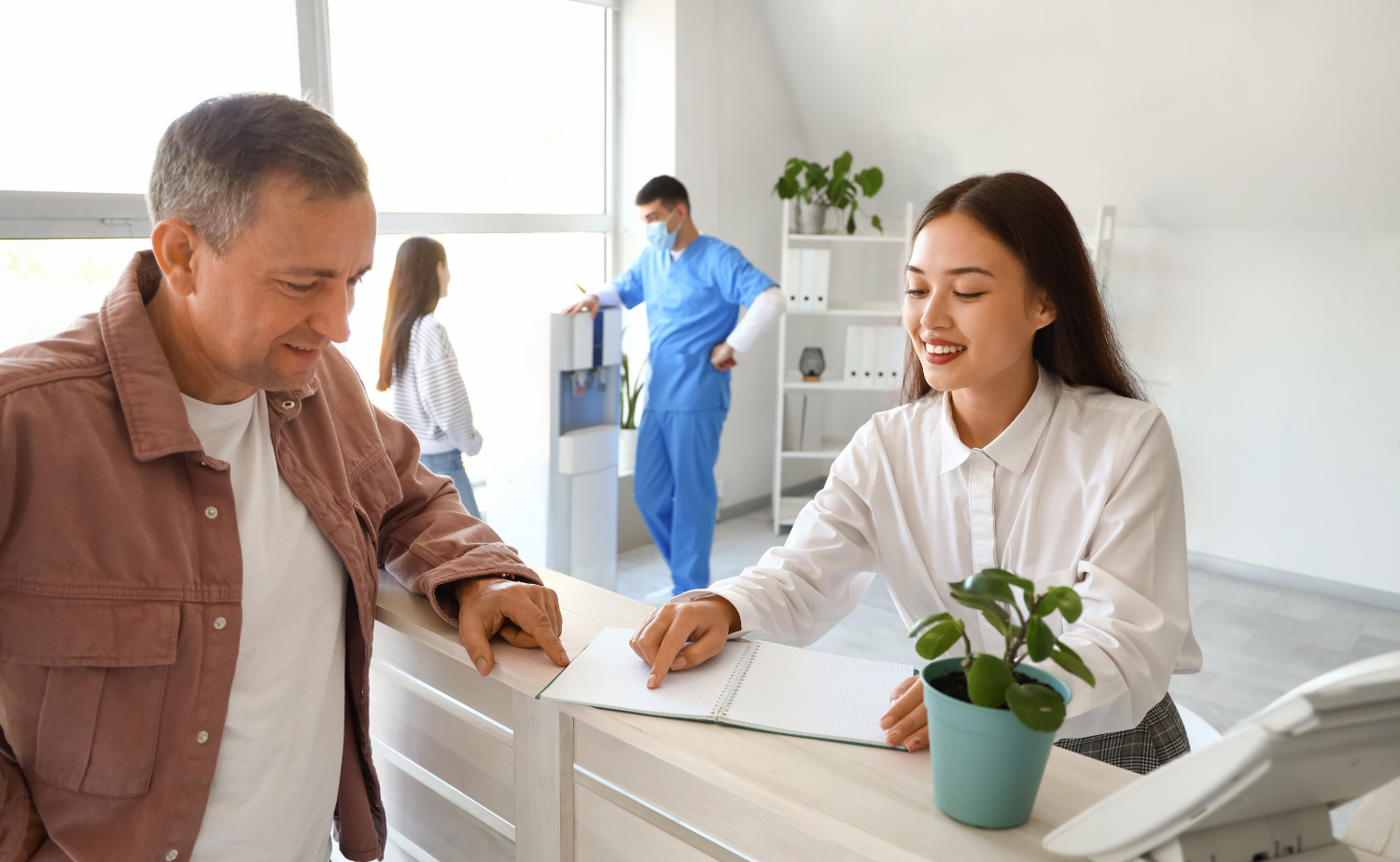  A cheerful receptionist assists a patient at a reception desk, with a healthcare professional and another patient in the background, creating a welcoming and efficient healthcare environment.