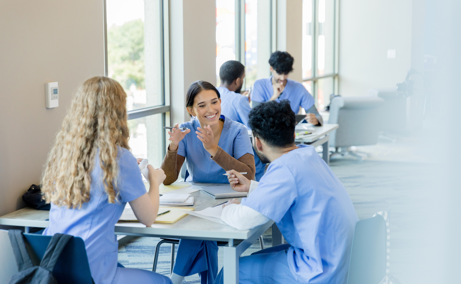 A group of healthcare students or professionals in blue scrubs are gathered around tables, engaged in animated discussion and collaboration, in a bright, modern setting with large windows.