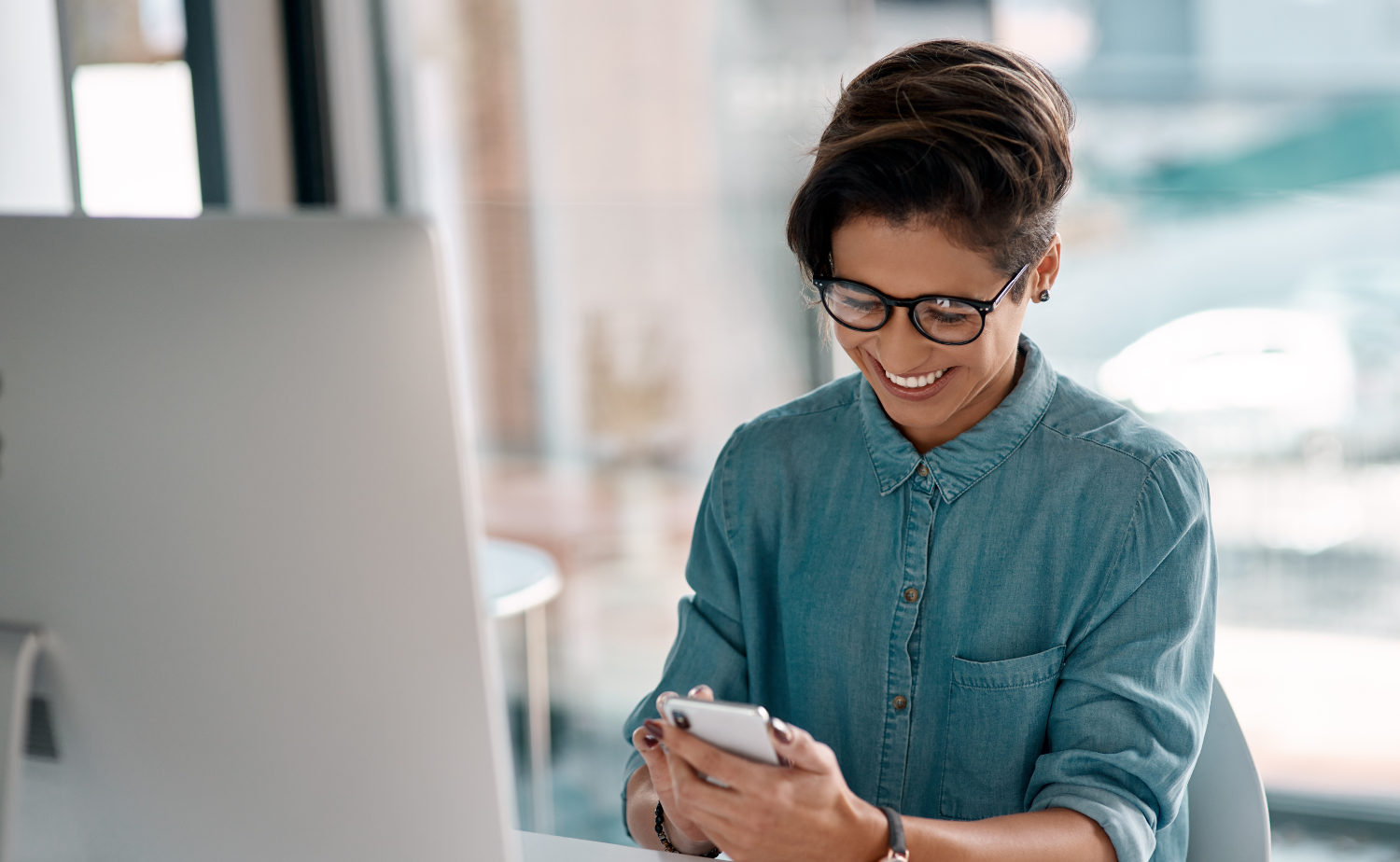 A person sitting at a desk and smiling while looking at their smartphone. They are wearing glasses and a denim shirt, with a computer monitor in front of them. The background shows a bright and modern office environment with large windows.