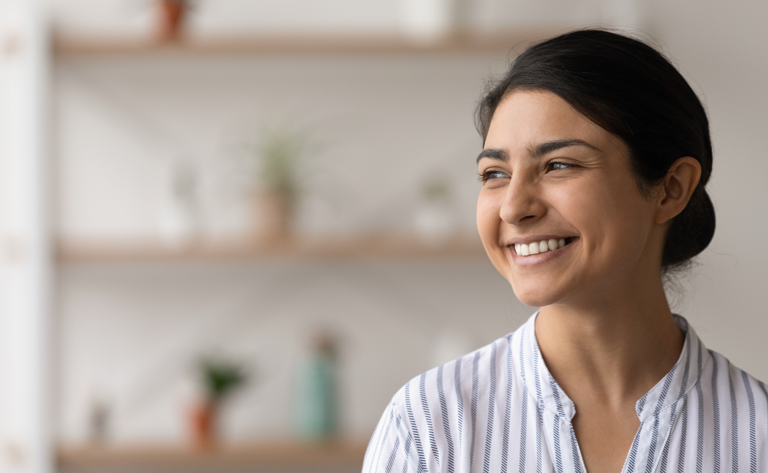 A young professional woman with dark hair smiles confidently while looking off to the side, set against a softly blurred background with shelves and plants, conveying positivity and optimism.