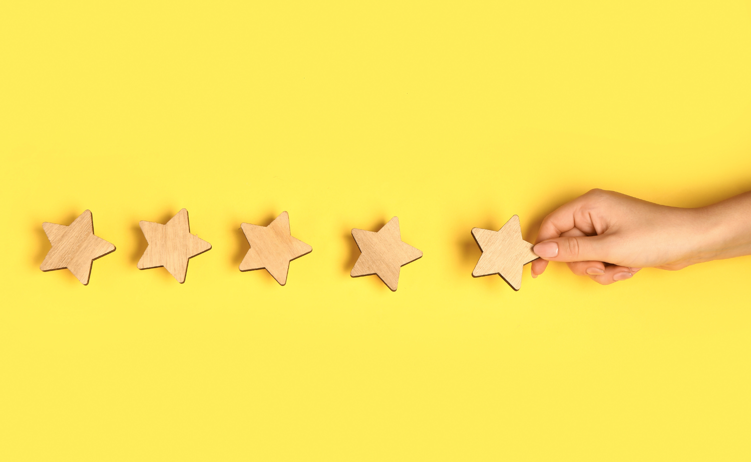 A hand places a wooden star among a row of four other stars on a bright yellow background, representing the concept of rating, review, or achieving a five-star quality.
