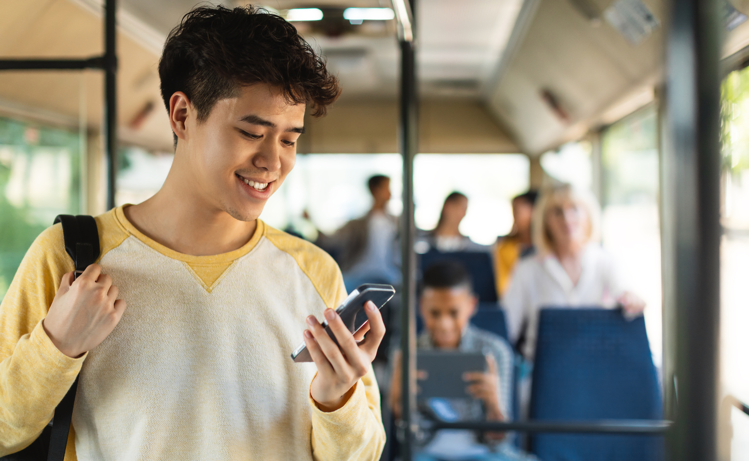 A young man in a yellow and white shirt smiles while looking at his phone on a bus, with other passengers engaged in their own activities in the background, representing connectivity and engagement.