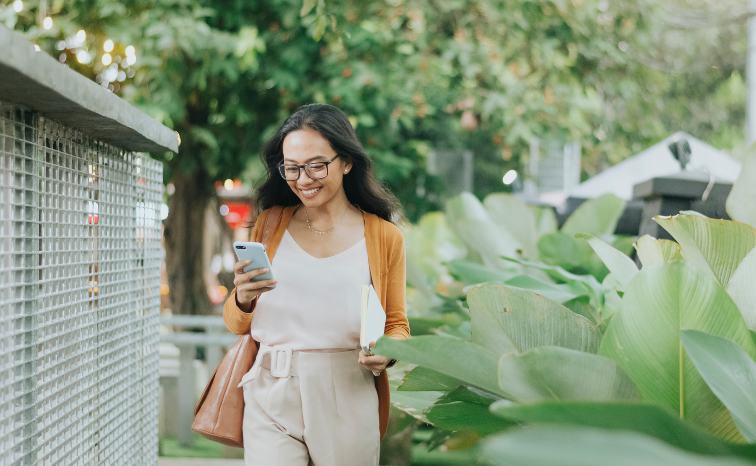A young woman walking outdoors and looking at her smartphone with a smile. She is wearing glasses, a white top, and a light brown cardigan, carrying a brown shoulder bag and a notebook. The background features lush green plants and a blurred urban setting.